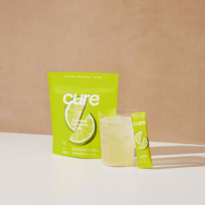 A bright green packet and a smaller sachet of Cure Hydrations Hydrating Electrolyte Drink Mix sit against a neutral background. In front, a glass with light yellow liquid and ice features Coconut Water and Pink Himalayan Salt.