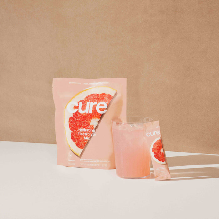 A package of Cure Hydrations Hydrating Electrolyte Drink Mix with grapefruit design sits next to a glass of pink beverage and a single-use sachet. Soft lighting on the neutral beige background and white surface enhances the drinks appeal.