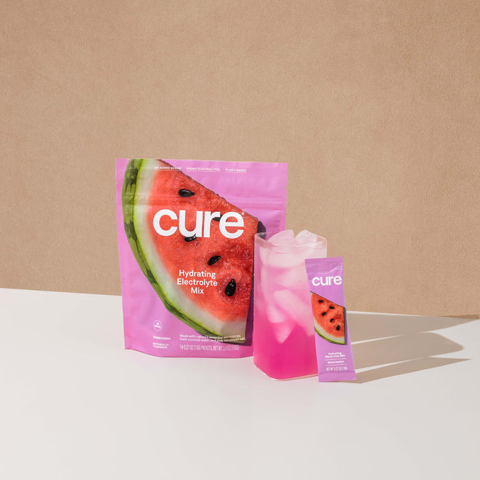 A pink pouch of Cure Hydrations Hydrating Electrolyte Drink Mix featuring a watermelon design is next to a clear glass of pink iced liquid. A small sachet lies on a light beige background, offering hydration similar to coconut water.