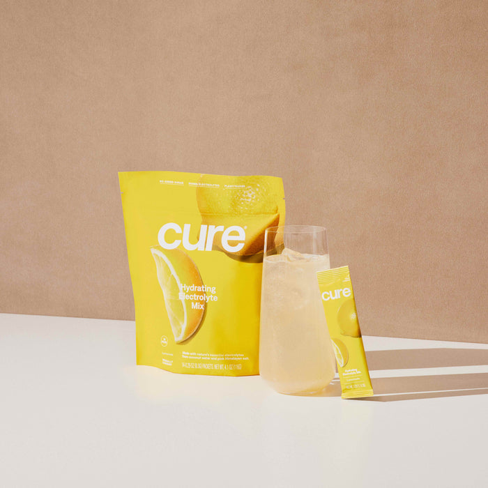 The yellow packaging of Cure Hydrations Hydrating Electrolyte Drink Mix, adorned with a lemon design, is displayed. Alongside it, a sachet and a glass of light beverage highlight quick absorption for effective hydration against a neutral background.