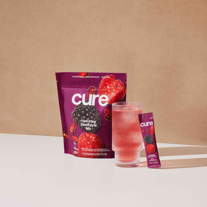 A package and sachet of Cure Hydrations Hydrating Electrolyte Drink Mix, designed for rapid absorption, accompany freeze-dried raspberries and blackberries next to a glass of pink liquid on a white surface against a brown background.