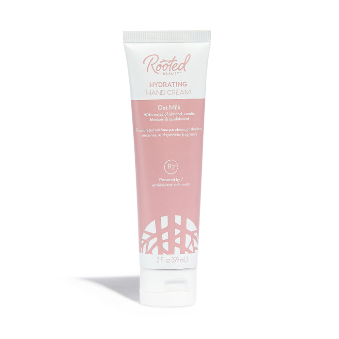 Rooted Beautys Hydrating Hand Cream in Oat Milk scent, enriched with plant-based moisturizers like shea butter, comes in a pink and white 2 fl oz (59 ml) tube that highlights ingredients and benefits. The packaging features a logo with interwoven lines at the bottom.