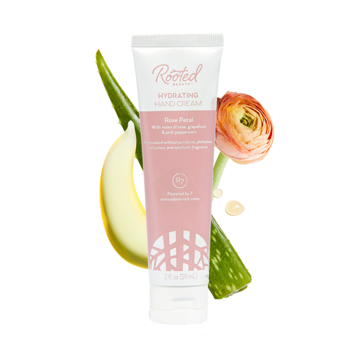 Rooted Beauty Hydrating Hand Cream in rose petal scent, enriched with shea butter, is placed alongside a pink flower, aloe vera leaf, and cream smear. This plant-based moisturizer offers dry skin relief and is packed with antioxidants, free of parabens and phthalates.