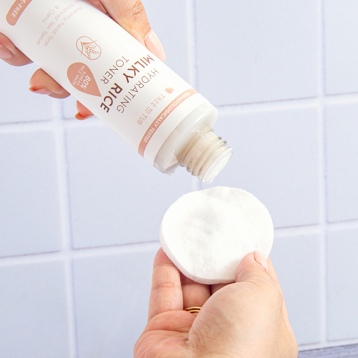 A person is pouring Tree To Tubs Hydrating Milky Rice Toner for Dry & Dehydrated Skin onto a white cotton pad from a bottle with a beige and white label featuring text and a logo, set against light blue tiles for a serene ambiance.