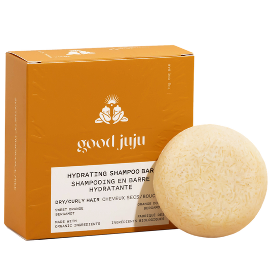 A round, beige organic Hydrating Shampoo Bar by Good Juju Body & Home sits in front of an orange box, offering frizz control for dry/curly hair with sweet orange and bergamot. Made with organic ingredients.