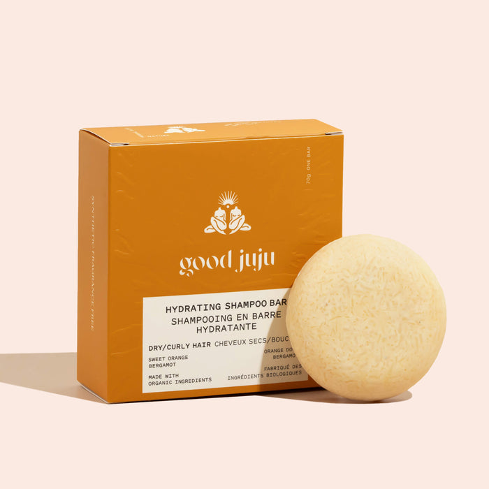 A round, beige Hydrating Shampoo Bar from Good Juju Body & Home sits next to an orange box highlighting frizz control and organic ingredients, labeled for dry/curly hair.
