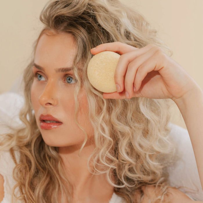 A woman with long, curly blonde hair holds the Hydrating Shampoo Bar by Good Juju Body & Home near her forehead, gazing thoughtfully to the side.