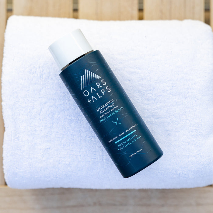 A bottle of Oars + Alps Hydrating Shampoo sits on a white folded towel on a wooden surface.