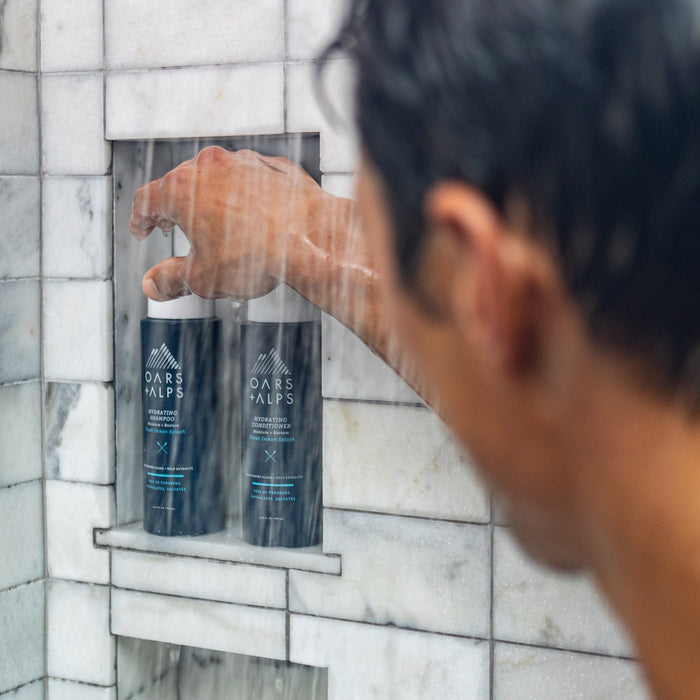From behind, a person reaches for Oars + Alps Hydrating Shampoo on a marble shower shelf with water running and another product nearby.