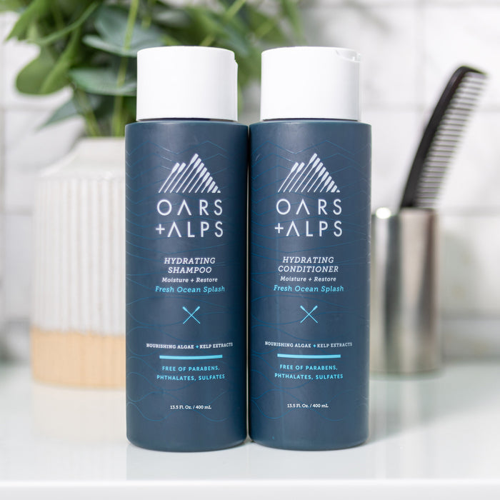 Two bottles of Oars + Alps Hydrating Shampoo are displayed side by side on a bathroom counter, with a toothbrush holder and green plant in the background, emphasizing their sulfate-free formula.