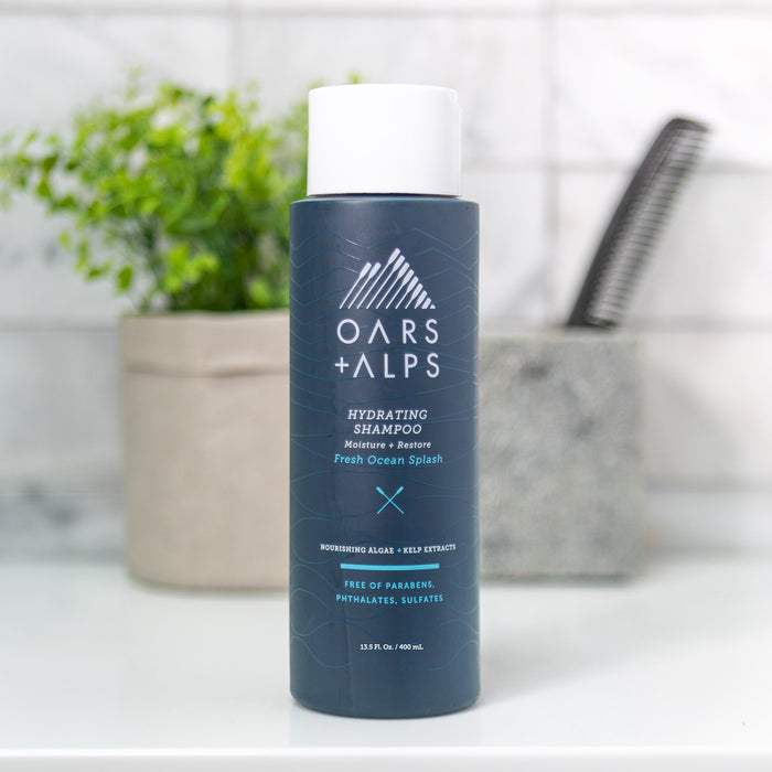 A dark blue Oars + Alps Hydrating Shampoo bottle sits on a bathroom counter beside a comb and gray towel. The label notes sea ingredients like aloe and kelp, and its free of parabens, phthalates, and sulfates.