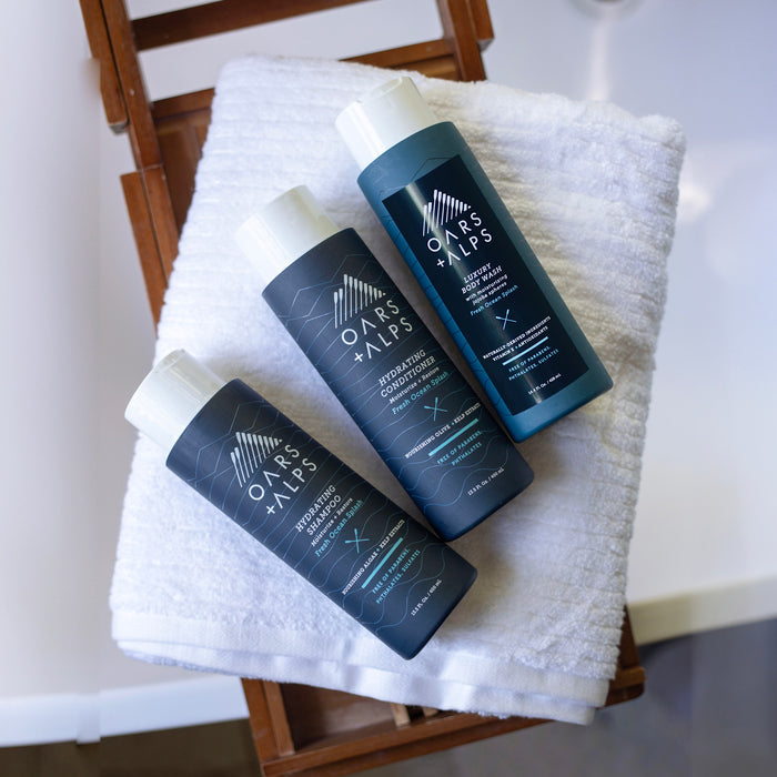 Oars + Alps Hydrating Shampoo and two other sulfate-free hair care bottles are displayed on folded white towels atop a wooden chair.