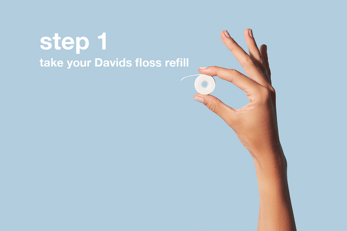 A hand holds a small spool of Davids Expanding Dental Floss with Hydroxyapatite against a light blue background. Text reads: step 1 take your Davids Expanding Dental Floss refill, part of our convenient refill system.