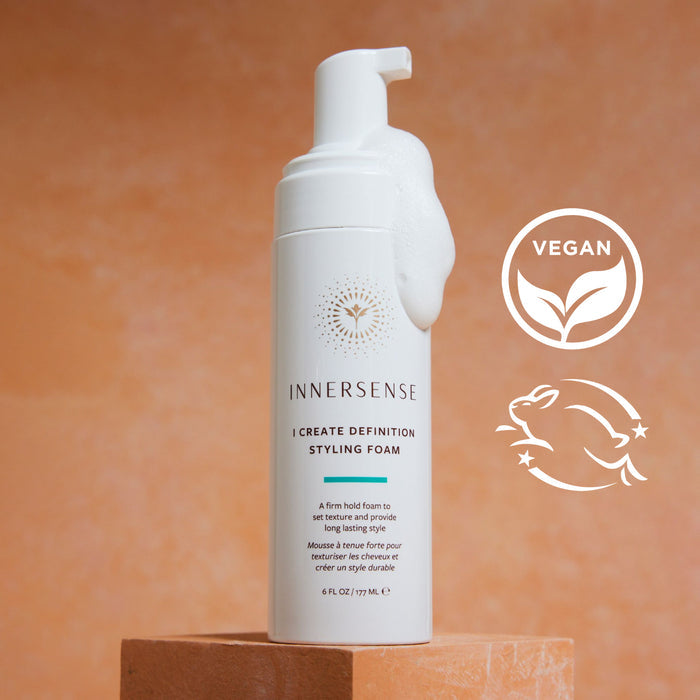 A bottle of Innersense I Create Definition Styling Foam on a textured orange surface. The bottle has foam on the dispenser and features vegan and eco-friendly icons. The background is a warm, earthy tone.