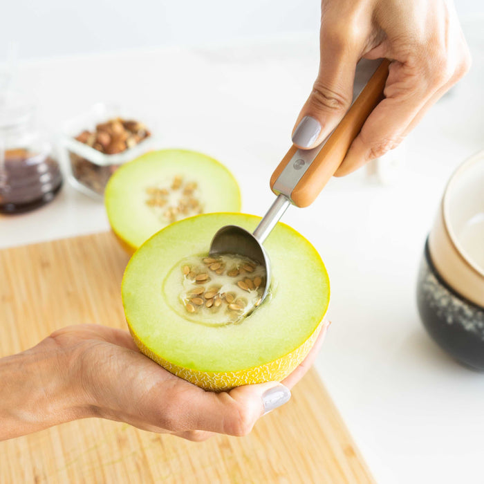 A person uses the Full Circle Ice Cream Scoop to remove seeds from half a yellow melon resting on their palm. A wooden cutting board and various small bowls are visible in the background. This stainless steel scoop is both sturdy and dishwasher safe for convenience.
