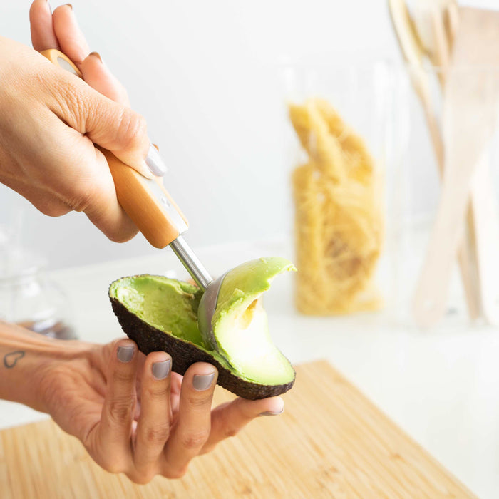 A person uses the Full Circle Ice Cream Scoop, featuring a wooden handle and stainless steel design, to remove avocado flesh from a halved avocado on a wooden cutting board. In the background, pasta and wooden utensils are visible.