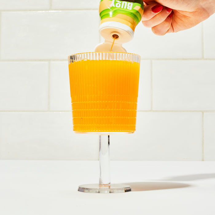 A clear, ribbed glass of bright orange juice on a white surface promises hydration and immunity with Buoy Hydrations Immunity Drops - Unsweetened Electrolytes + Immune Support for Any Beverage being poured from a green and white bottle. The backdrop showcases white tiled walls.