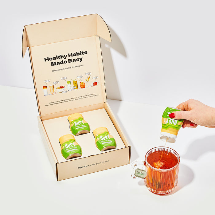 A hand squeezes a green Buoy Hydration Immunity Drops bottle, featuring electrolytes and immune support, into a glass mug next to an open box with more bottles. The lid reads Healthy Habits Made Easy.