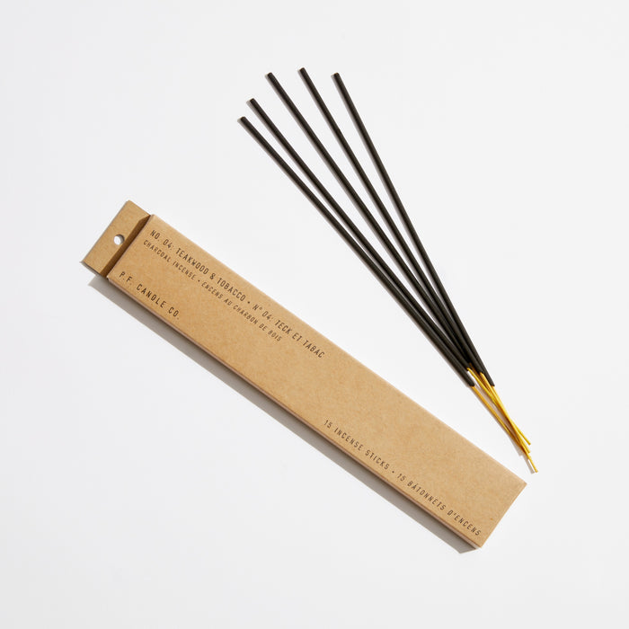 A brown P.F. Candle Co. Teakwood & Tobacco box lies open on a white surface, revealing the signature scents of teakwood and sandalwood. Four black Incense Sticks with golden ends rest beside it, inviting an aromatic experience.