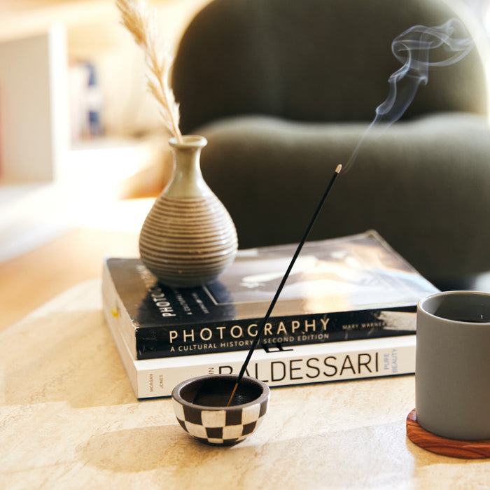 A cozy scene includes a table with a lit P.F. Candle Co. sandalwood rose incense stick, a checkered holder, a vase of dried grasses, stacked photography books, and a gray mug. Soft lighting engulfs the area in warmth, with a blurred green chair visible in the background.