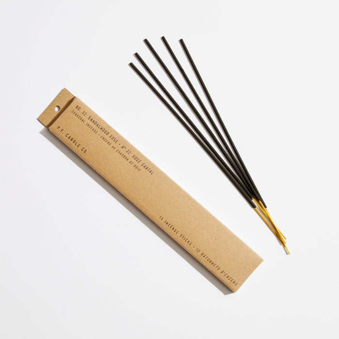 Brown P.F. Candle Co. packaging sits next to four long, thin black Incense Sticks with yellow tips, likely sandalwood-scented. The sticks fan out on a white background, exuding signature scents.