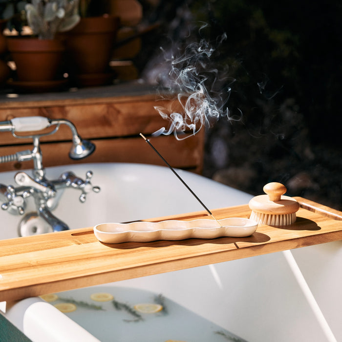 A P.F. Candle Co. Incense Stick burns on a wooden bath tray, with its sandalwood scent mingling with the steam. Nearby, a soap scrub brush rests in a ceramic holder above a white bathtub filled with water and floating lemon slices amid potted plants adding greenery.