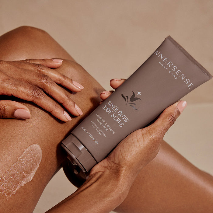 A hand holds a tube of Inner Glow Body Scrub by Innersense Organic Beauty against a leg, featuring its gel formula with a small applied smear. The gray tube with white text emphasizes the routines focus on physical exfoliators for improved skincare outcomes.