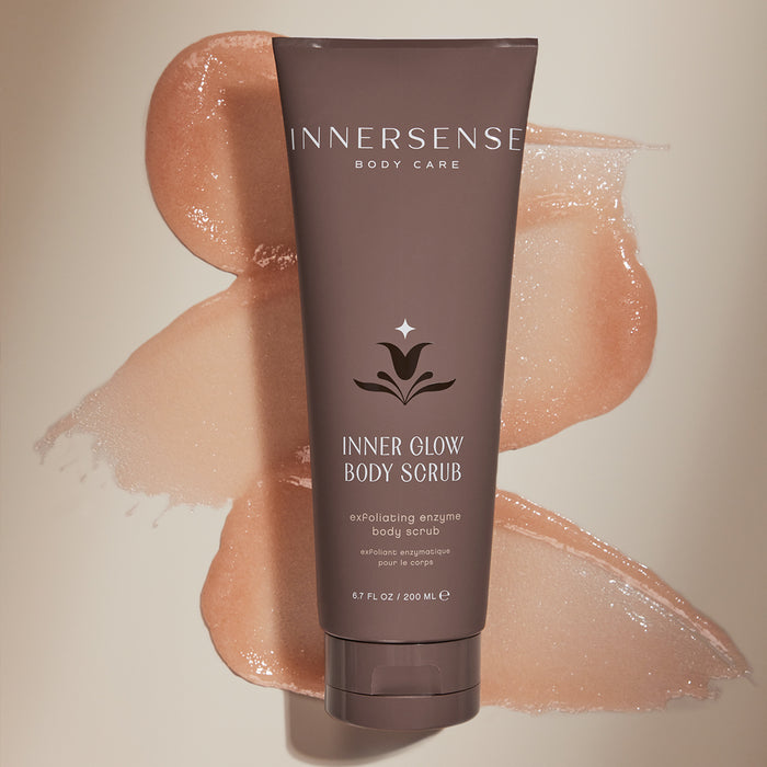 A tube of Innersense Organic Beauty Inner Glow Body Scrub stands on a beige background, with its creamy texture artfully spread behind it. The text promises your healthiest skin yet with powerful exfoliators, describing it as an exfoliating enzyme body scrub.
