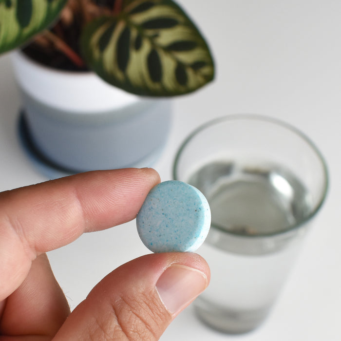 A hand holds an Instant Plant Food Tablet above a white surface, preparing to nourish houseplants. In the background, theres a glass of water and a potted plant with patterned green leaves thriving indoors.