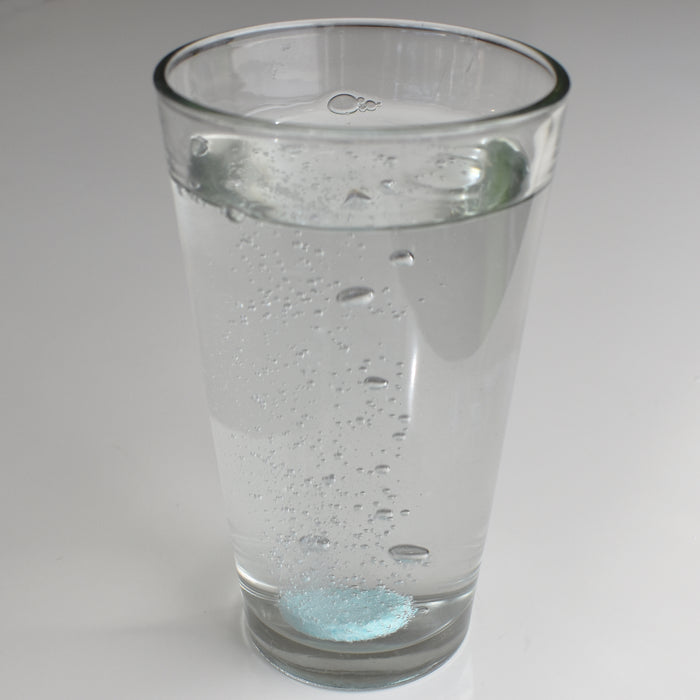 A clear glass of water holds a light blue Instant Plant Food Tablet dissolving at the bottom, releasing bubbles that rise to the surface. This invigorating sight mirrors how Instant Plant Food energizes your indoor plants, all against a plain white background.