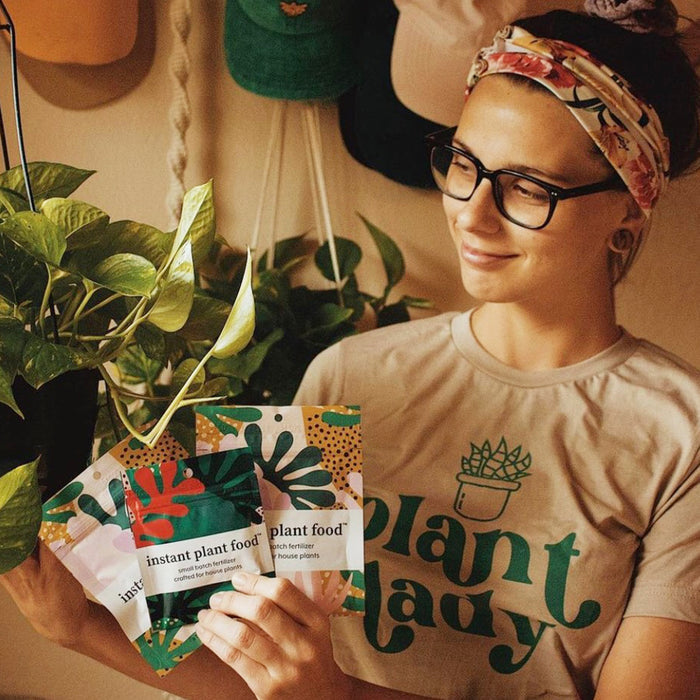 A person wearing glasses and a floral headband smiles while holding Instant Plant Food Tablets. Their plant lady T-shirt complements the lush indoor plants nearby. In the background, hats hang gracefully, completing this charming botanical scene.