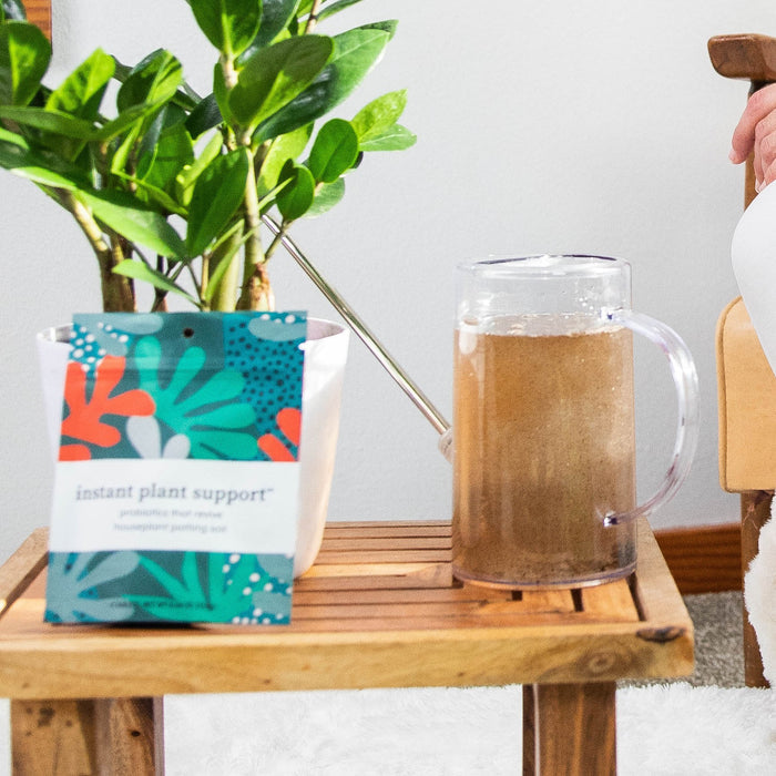 A packet labeled Instant Plant Food: Instant Plant Support sits on a wooden table beside a pitcher of brown liquid. In the background, a thriving green plant suggests magic beneath the surface with houseplant probiotics nurturing healthy soil.