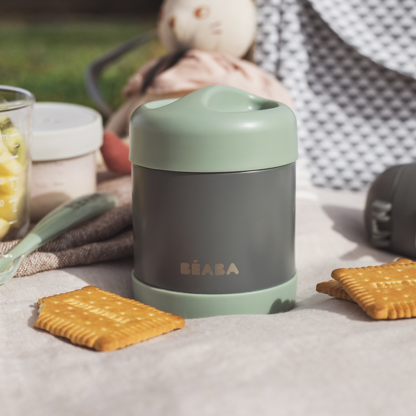 A gray and mint green BEABA Insulated Stainless Steel, Kid Friendly Food Container Jar sits on a picnic blanket with square biscuits and a baby food jar. A blurred stuffed toy enhances the cozy outdoor setting.