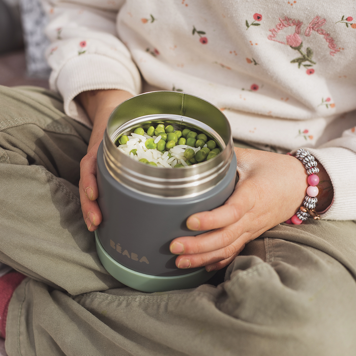 A child, dressed in a floral-patterned sweater and khaki pants, with beaded bracelets on one wrist, sits cross-legged holding a BEABA Insulated Stainless Steel, Kid Friendly Food Container Jar in green and gray, enjoying rice and peas from the leak-proof jar.