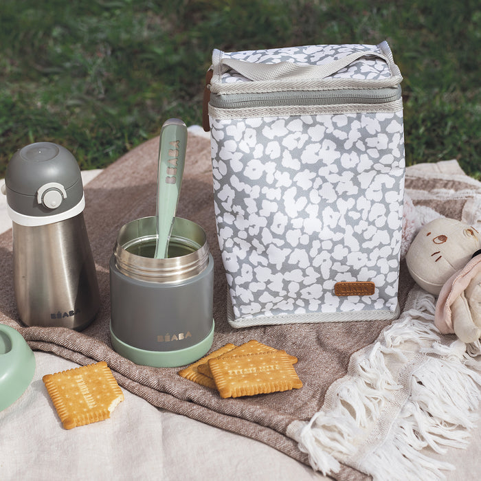 A summer picnic setup includes a patterned lunch bag, the BEABA Insulated Stainless Steel Food Container Jar, a leak-proof container with a green spoon, and scattered crackers. A plush toy and a white fringed blanket add to the cozy outdoor scene on the grass.