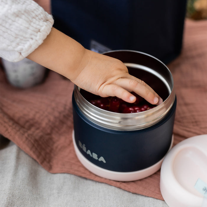 A childs hand reaches into BEABAs Insulated Stainless Steel, Kid Friendly Food Container Jar in navy blue, filled with cherries. The leakproof jar rests on a soft pink cloth beside another container and a dark bag, creating a charming picnic scene.