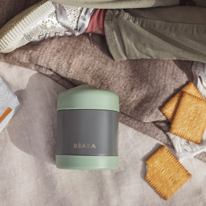A BEABA Insulated Stainless Steel, Kid Friendly Food Container Jar in green and gray rests on a picnic blanket next to two crackers. In the background, a persons leg wearing khaki pants and textured sneakers can be seen.