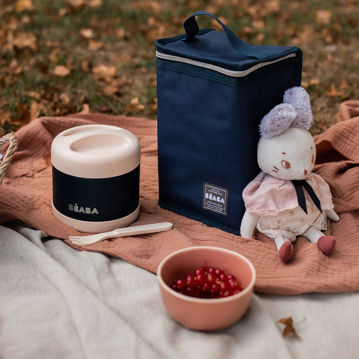 A picnic on a blanket includes a navy and cream thermos, plush doll, navy insulated bag, and a pink bowl with red currants accompanied by BEABAs Insulated Stainless Steel Kid Friendly Food Container Jar for temperature-controlled meals. Scattered leaves dot the grassy background.