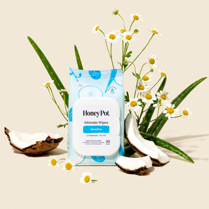 A pack of The Honeypot Company Intimate Wipes - Sensitive is displayed on a beige background, surrounded by aloe vera leaves, chamomile flowers, and cracked coconut pieces.