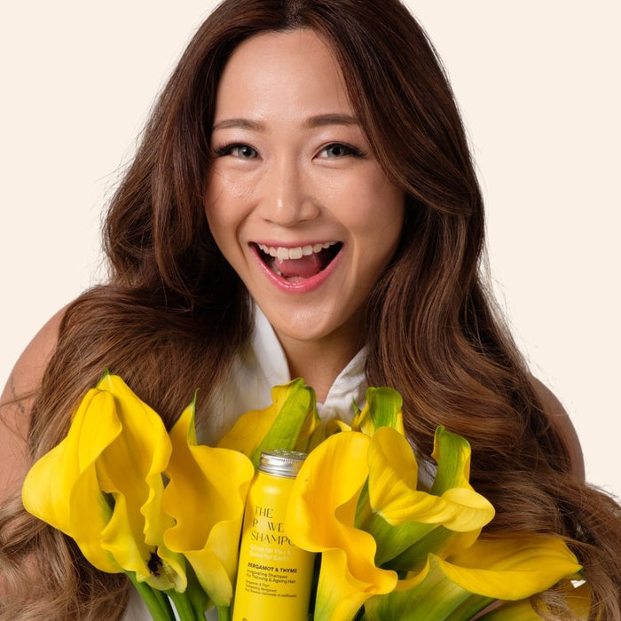 A smiling woman with long brown hair holds a bouquet of vibrant yellow flowers and a bottle labeled Invigorating & Stimulating Powder Shampoo by The Powder Shampoo, set against an off-white background, highlighting her joyful expression and hinting at enhanced hair growth potential.