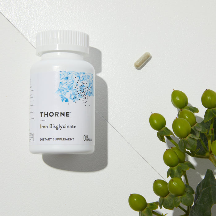 A Thorne Iron Bisglycinate dietary supplement bottle is placed on a white surface, supporting healthy red blood cells. Nearby, a single capsule rests alongside green berries and leaves partially visible in the bottom right corner.