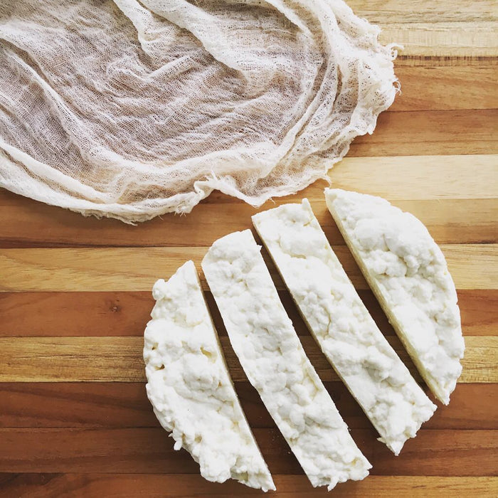Five wedges made using the FarmSteady Italian Fresh Cheese Making Kit rest on a wooden cutting board beside cheesecloth, featuring a crumbly, slightly uneven texture.