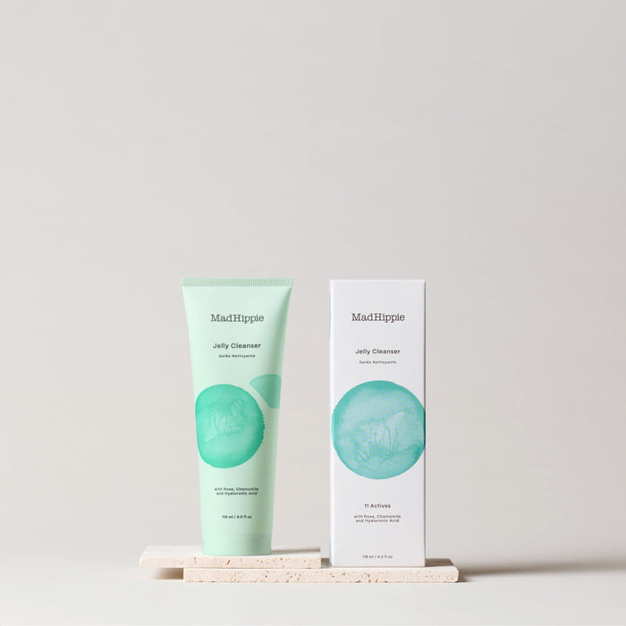 A green Mad Hippie Jelly Cleanser tube with antioxidant benefits stands beside its matching box on a light stone surface, both showcasing minimalist designs with the brands name and a circular blue-green graphic on an off-white background.