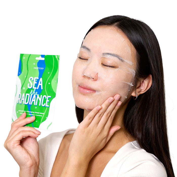A long-haired individual wears a translucent K-beauty face mask from the K Beauty Face Mask Set by FaceTory, gently touching their face while holding a colorful Sea the Radiance packet. The background is white.