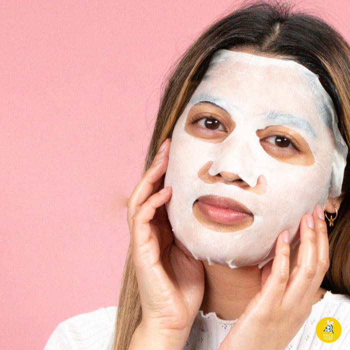 A person with long hair enjoys a FaceTory K Beauty Face Mask ritual, gently touching the essence-soaked mask. A solid pink background highlights this self-care must-have moment.