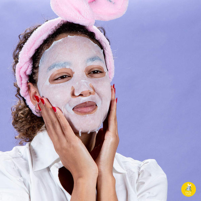 Dressed in a white shirt, someone pampers themselves with the FaceTory K Beauty Face Mask and a pink bunny-ear headband, gently touching their cheeks. Against a light purple background, their nails are painted red. A small yellow logo appears in the bottom right corner.