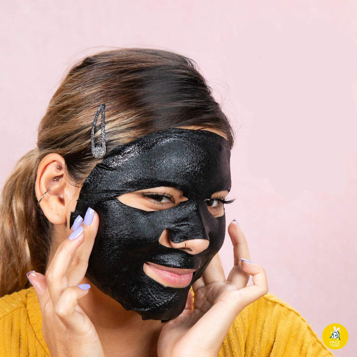 A person with light brown hair, in a yellow top, wears a black FaceTory K Beauty Face Mask from the FaceTory set, a self-care must-have. They adjust it with painted nails against a soft pink background.