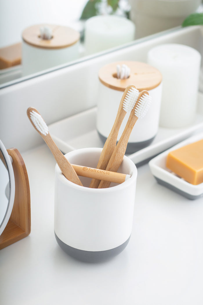 Three wooden toothbrushes sit in a Full Circle Keep It Clean Toothbrush Holder on a bathroom counter. Nearby, theres a bar of soap in a dish and a white container. A mirror reflects the tranquil scene, embodying Dry Earth™s simplicity and sustainability theme.