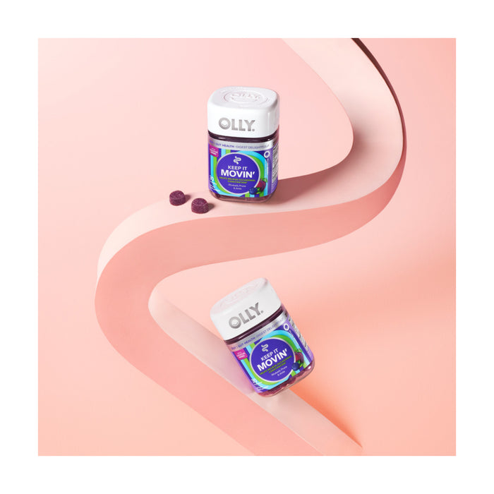 Two OLLY Keep It Movin vitamin gummy containers rest on a curvy pink surface. The jars are surrounded by scattered gummies to promote digestive health, with the soothing pink backdrop enhancing the wellness theme.