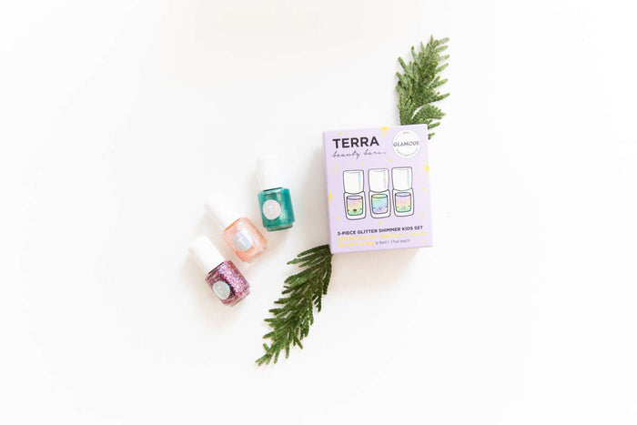 Three bottles of glittery nail polish in pink, teal, and purple are placed beside a box labeled Terra Beauty Bars featuring the Kids 3-Piece Glitter Shimmer Nail Collection, showcasing this cruelty-free set on a white background with green sprigs.